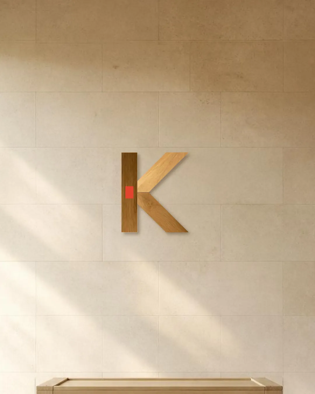 The two-part K as an oak relief on a limestone wall in raking window light, its joint showing red.